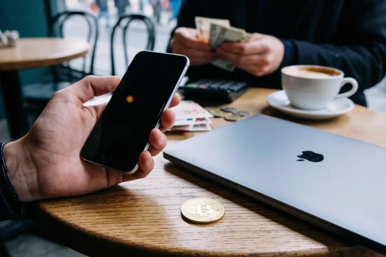 Main tenant un smartphone, ordinateur portable, pièce Bitcoin, café et billets sur une table en bois.