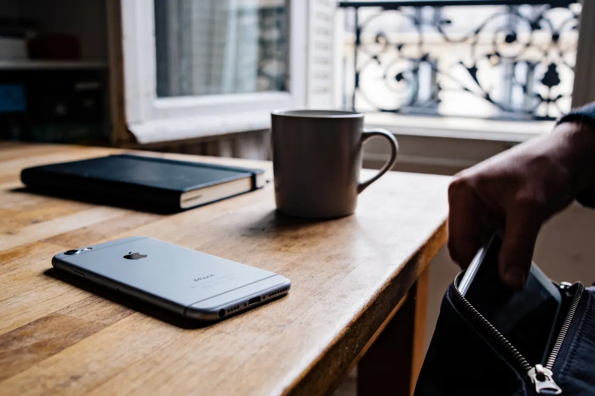 Smartphone sur table en bois, carnet, tasse, main tenant une tablette près d'une fenêtre ouverte.