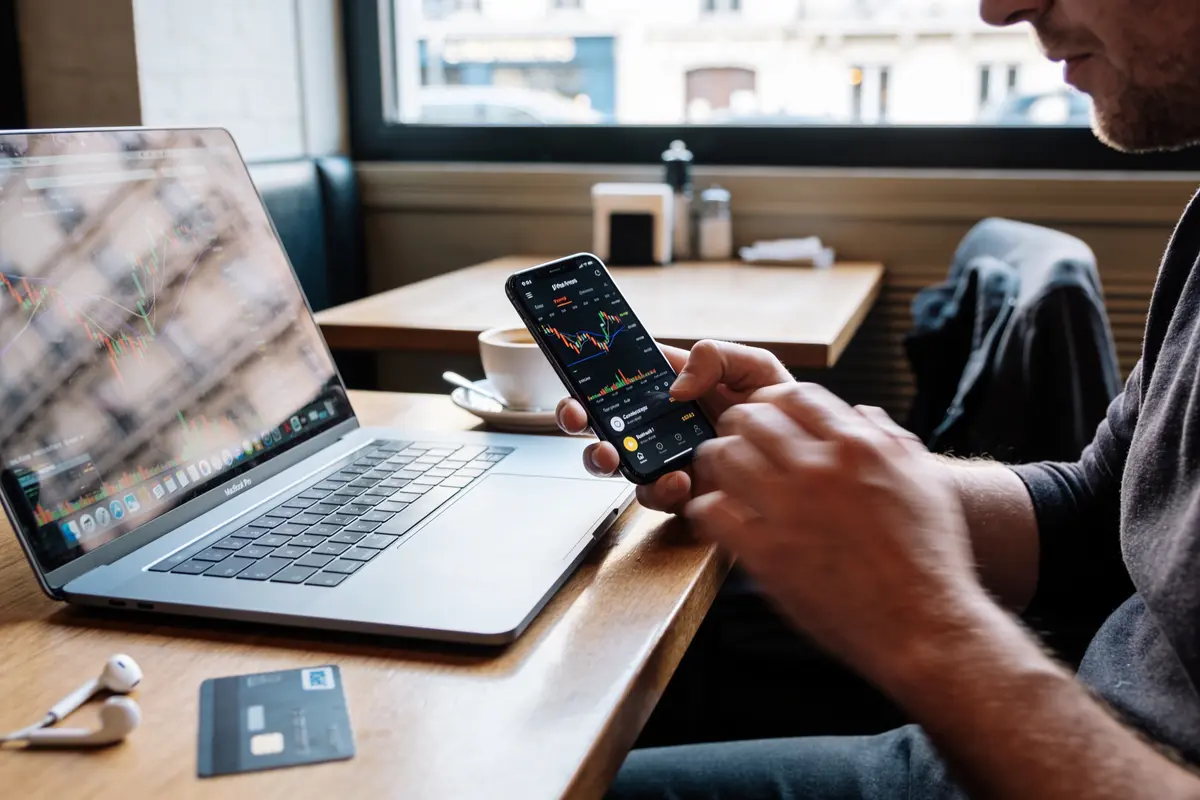 Homme utilisant un smartphone et un ordinateur portable avec graphiques financiers, café, écouteurs et carte bancaire sur la table.