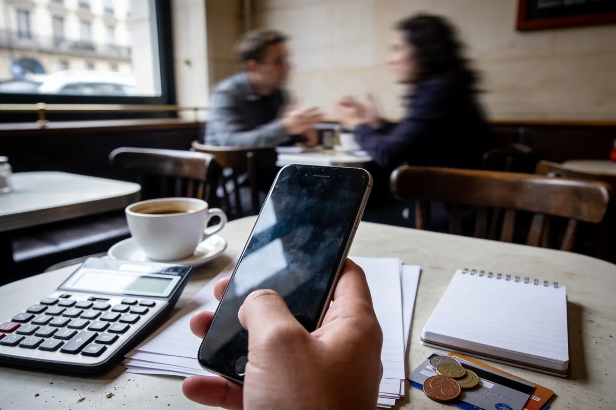Main tenant un smartphone, café, calculatrice, bloc-notes, cartes bancaires sur table de café. Deux personnes floues discutent.